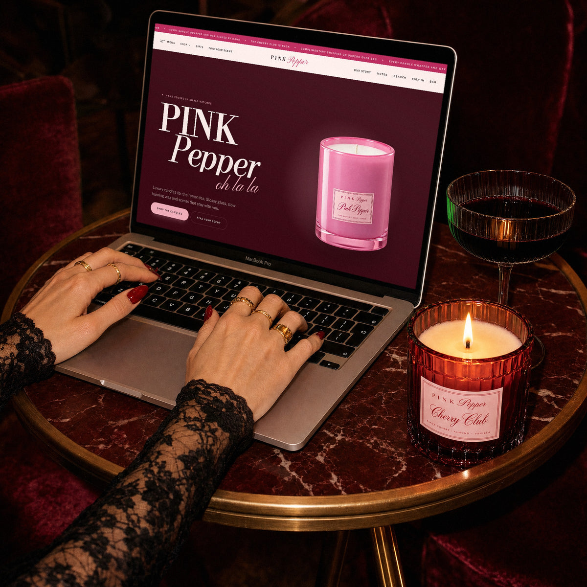Pink Pepper World homepage on a laptop, hands in black lace typing beside a lit Cherry Club candle